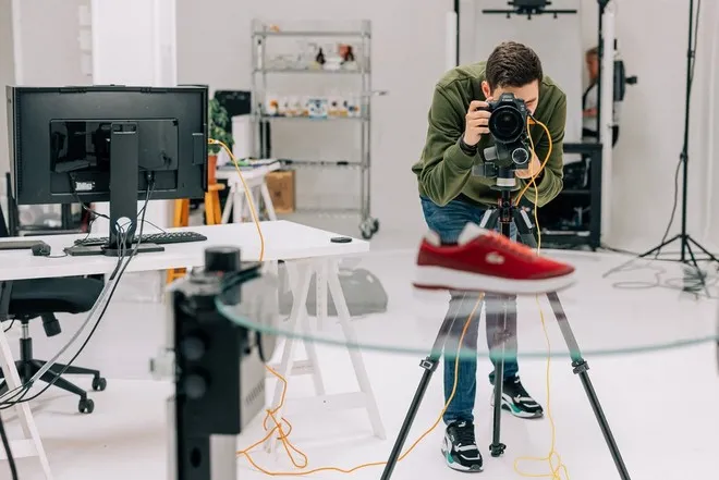 A man in a photography studio adjusts a Canon EOS camera pointing at a red trainer on a glass table in the foreground.