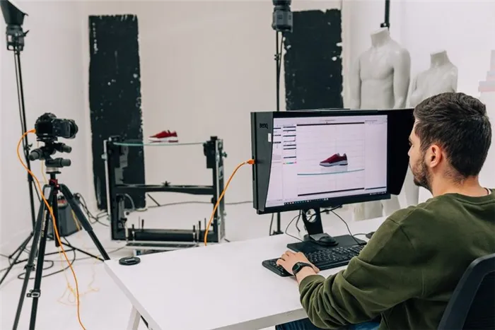 Камера Canon EOS, стоящая на штативе в студии, направлена на стеклянный стол, на котором стоит один красный кроссовок. Мужчина просматривает изображение кроссовка на ПО, открытом на мониторе.