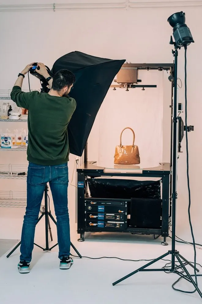 A man adjusts a studio light shining on a handbag on a mechanised display stand.